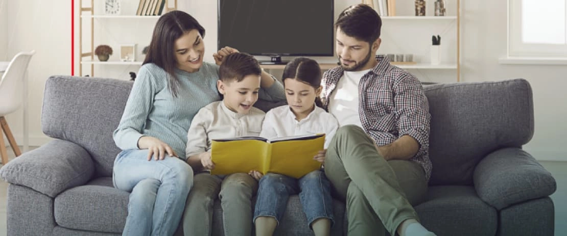Uma família de quatro pessoas está sentada em um sofá. Há uma mulher adulta, um homem adulto e um casal de crianças, com as crianças sentadas no meio entre os dois adultos. As crianças seguram um livro, e toda a família está olhando para o livro com expressões felizes. Ao fundo, há uma estante e uma TV