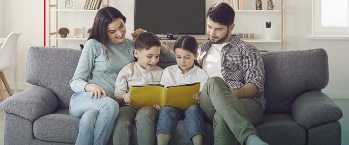 Uma família de quatro pessoas está sentada em um sofá. Há uma mulher adulta, um homem adulto e um casal de crianças, com as crianças sentadas no meio entre os dois adultos. As crianças seguram um livro, e toda a família está olhando para o livro com expressões felizes. Ao fundo, há uma estante e uma TV